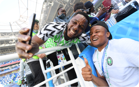 Two Nigerian football fans taking a selfie near stadium railings during a match.