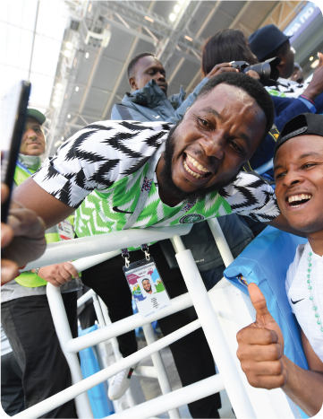 Two Nigerian football fans taking a selfie near stadium railings during a match.